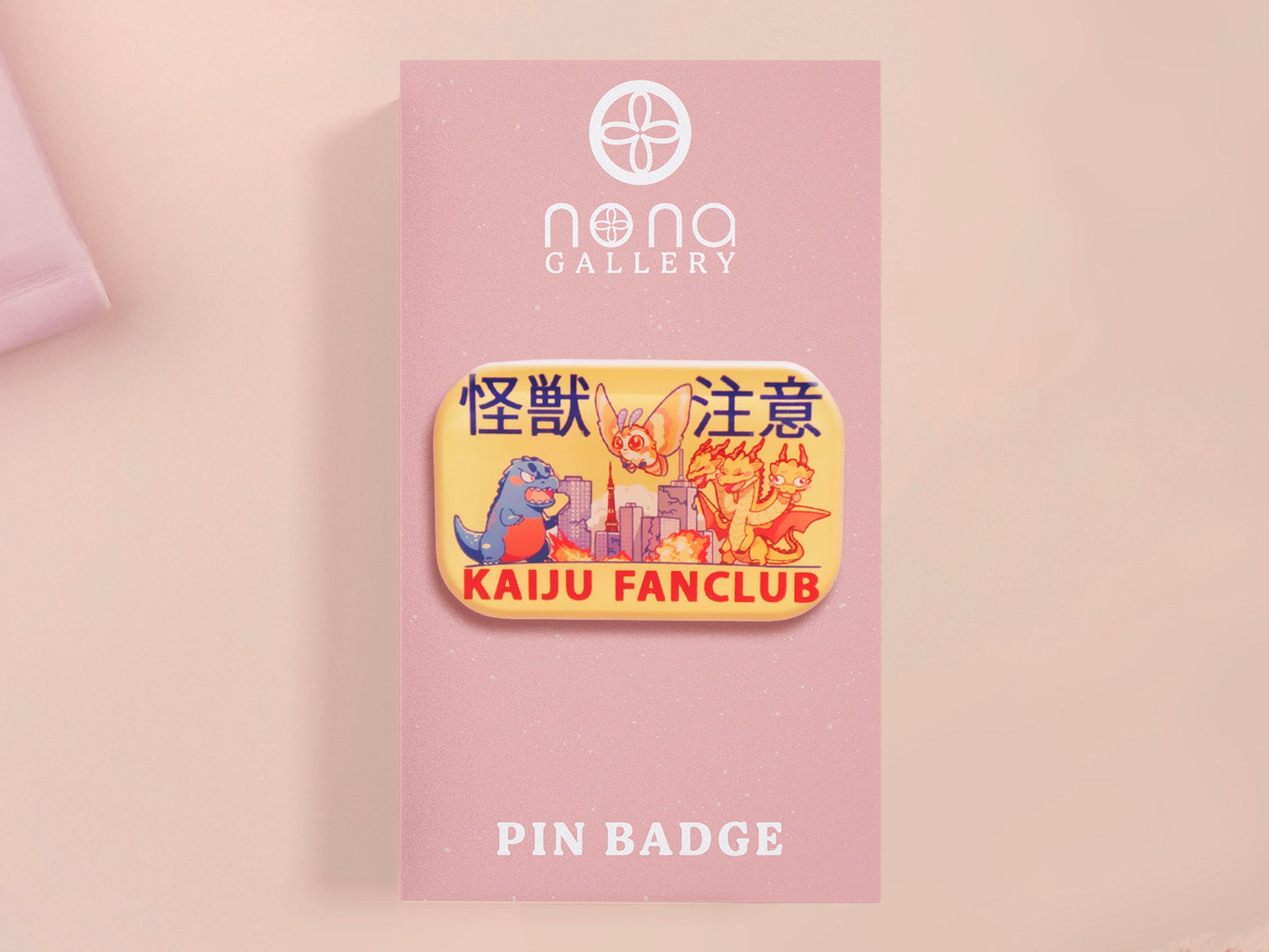 A clear acrylic pin badge with a pink heart-shaped pin back clasp. The design of the pin badge is of a cute chibi blue godzilla like kaiju mothra and King Ghidorah destroying a city with the quote kaiju fanclub and kaiju warning in japanese kanji