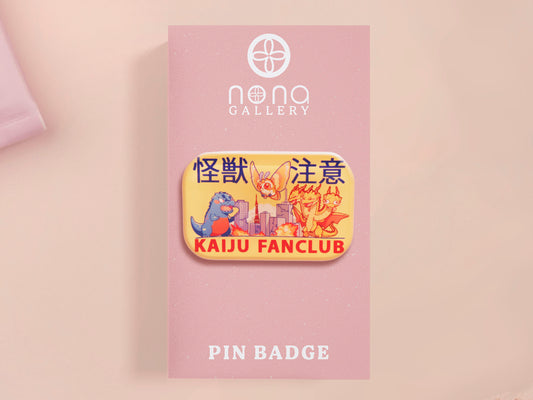 A clear acrylic pin badge with a pink heart-shaped pin back clasp. The design of the pin badge is of a cute chibi blue godzilla like kaiju mothra and King Ghidorah destroying a city with the quote kaiju fanclub and kaiju warning in japanese kanji