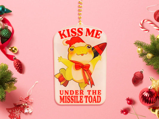 Acrylic epoxy christmas tree hanging decoration with a large acrylic charm of a cute chibi santa frog with a missile tied to its back with a red ribbon with the quote kiss me under the missile toad