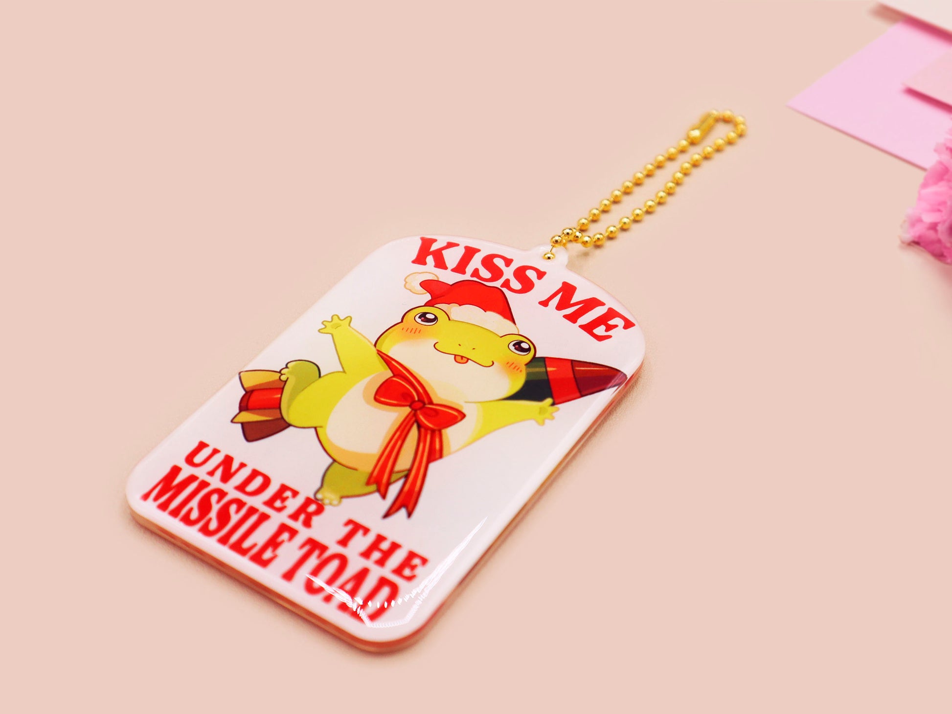 Acrylic epoxy christmas tree hanging decoration with a large acrylic charm of a cute chibi santa frog with a missile tied to its back with a red ribbon with the quote kiss me under the missile toad