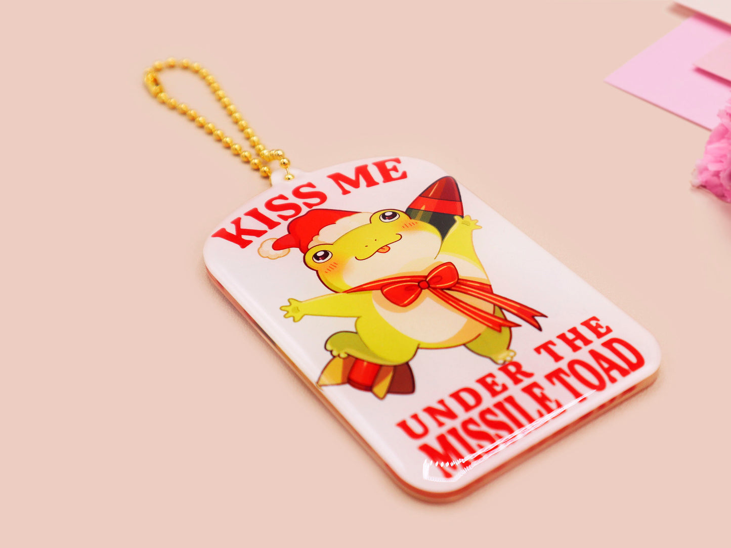 Acrylic epoxy christmas tree hanging decoration with a large acrylic charm of a cute chibi santa frog with a missile tied to its back with a red ribbon with the quote kiss me under the missile toad