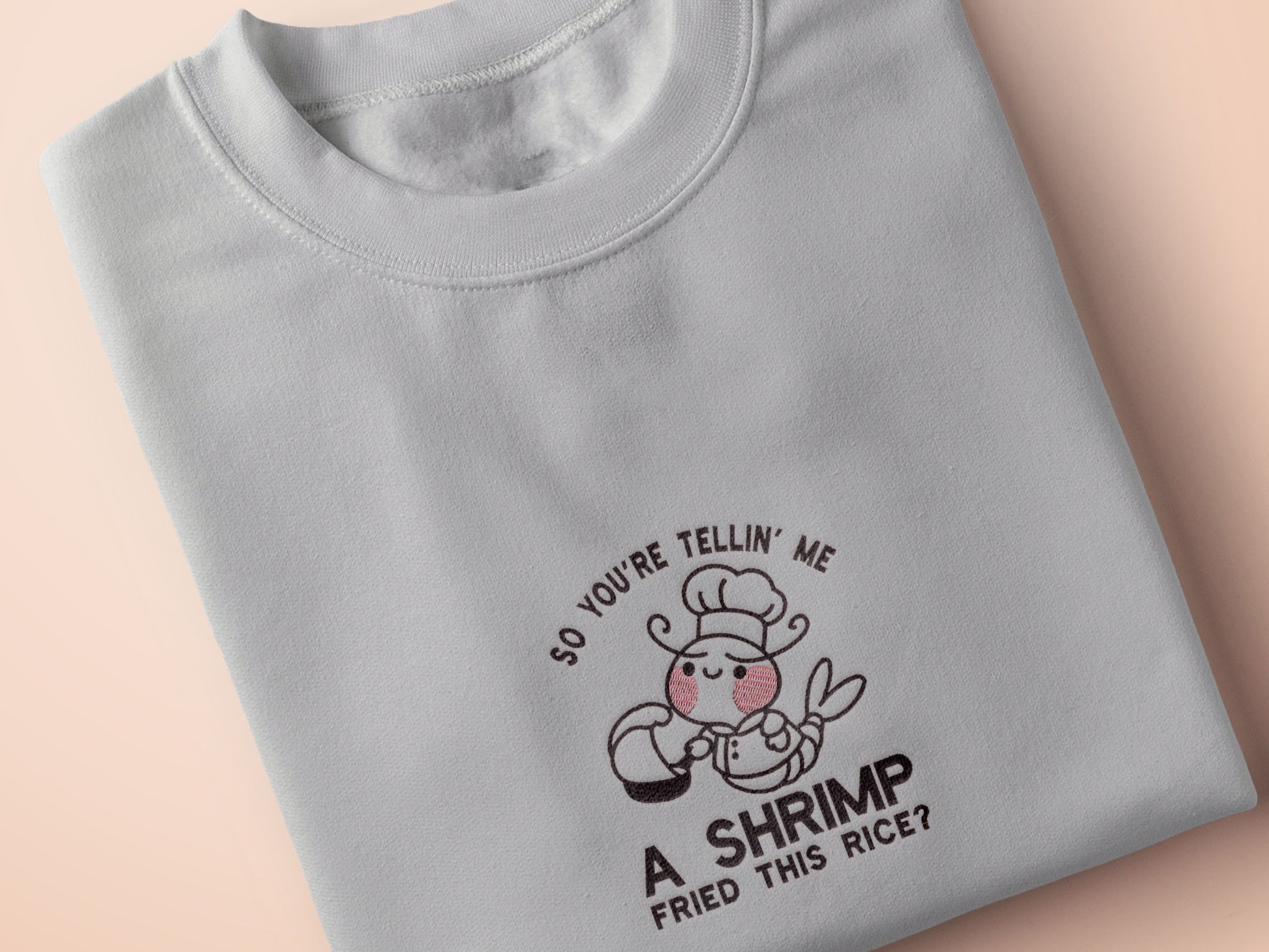 A grey embroidered t-shirt design of a cute chibi shrimp with pink cheeks dressed as a chef using a frying pan surrounded by the quote So You're Telling Me A Shrimp Fried This Rice?
