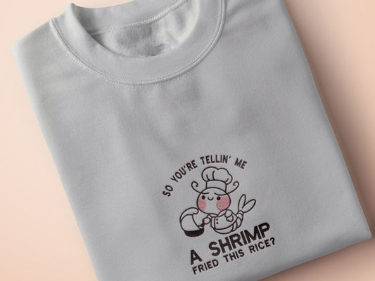 A grey embroidered t-shirt design of a cute chibi shrimp with pink cheeks dressed as a chef using a frying pan surrounded by the quote So You're Telling Me A Shrimp Fried This Rice?