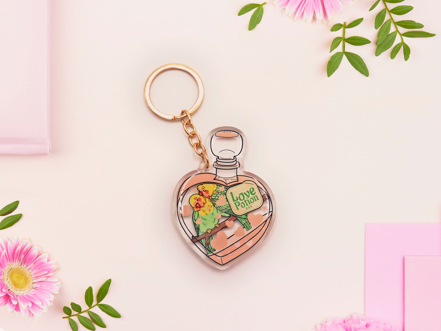 Clear acrylic double sided keyring with gold clasp, with a cute cartoon illustrated design of a heart shaped potion bottle containing two green and yellow love birds kissing each other labelled love potion xxx