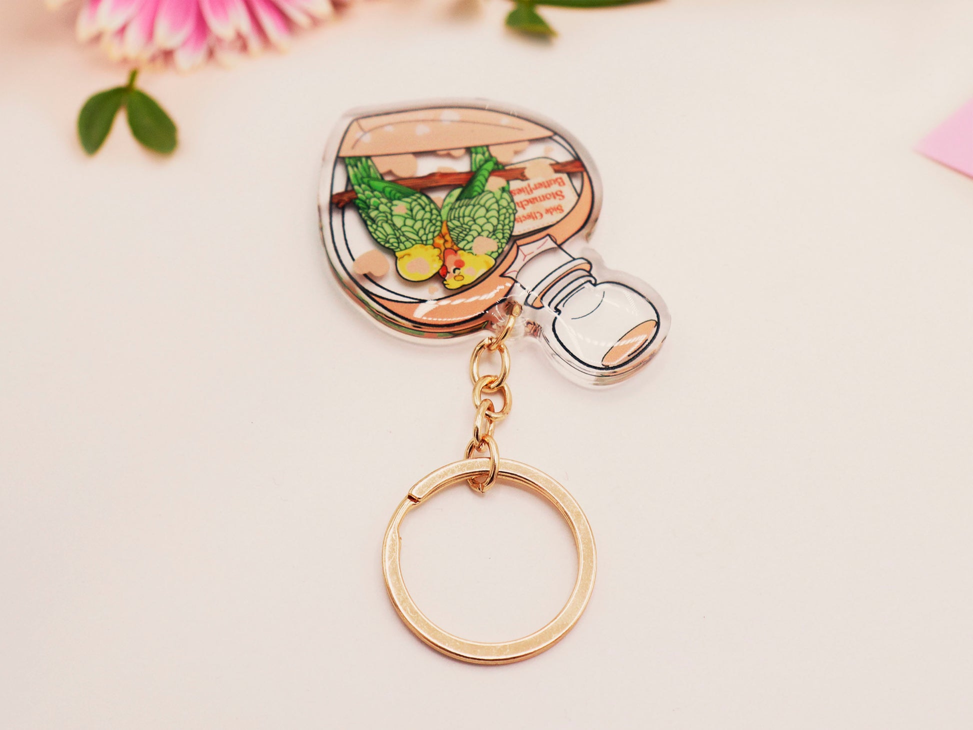 Clear acrylic double sided keyring with gold clasp, with a cute cartoon illustrated design of a heart shaped potion bottle containing two green and yellow love birds kissing each other labelled love potion xxx