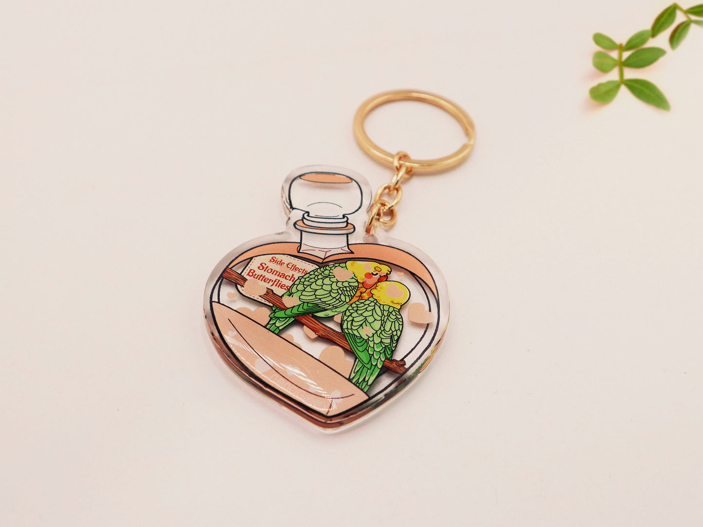 Clear acrylic double sided keyring with gold clasp, with a cute cartoon illustrated design of a heart shaped potion bottle containing two green and yellow love birds kissing each other labelled love potion xxx