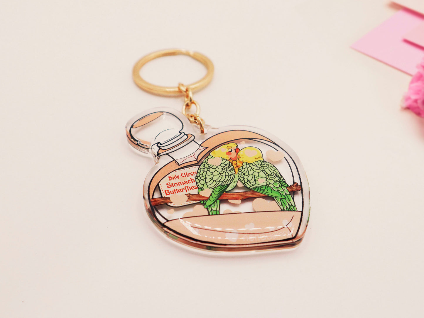 Clear acrylic double sided keyring with gold clasp, with a cute cartoon illustrated design of a heart shaped potion bottle containing two green and yellow love birds kissing each other labelled love potion xxx