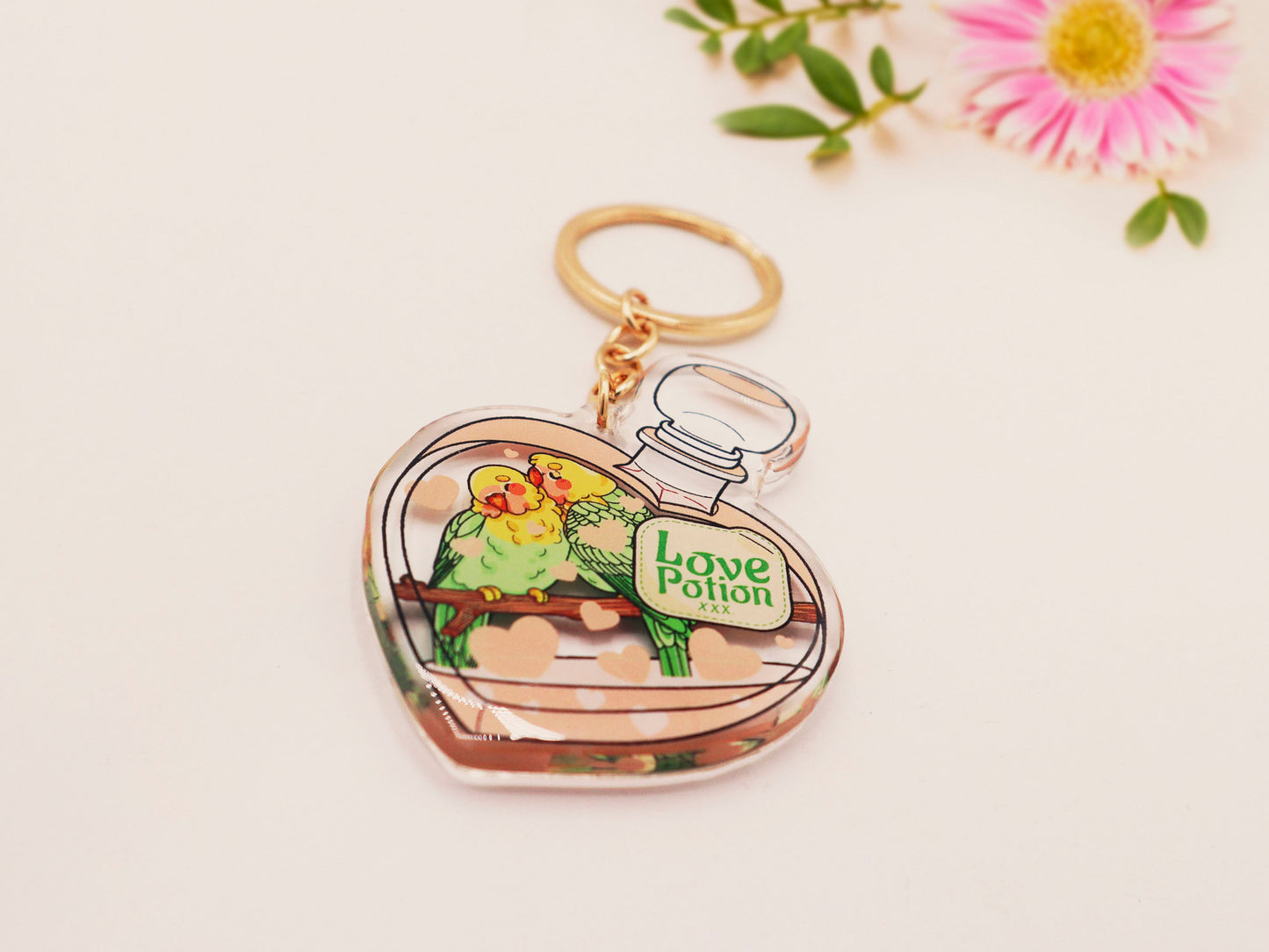 Clear acrylic double sided keyring with gold clasp, with a cute cartoon illustrated design of a heart shaped potion bottle containing two green and yellow love birds kissing each other labelled love potion xxx