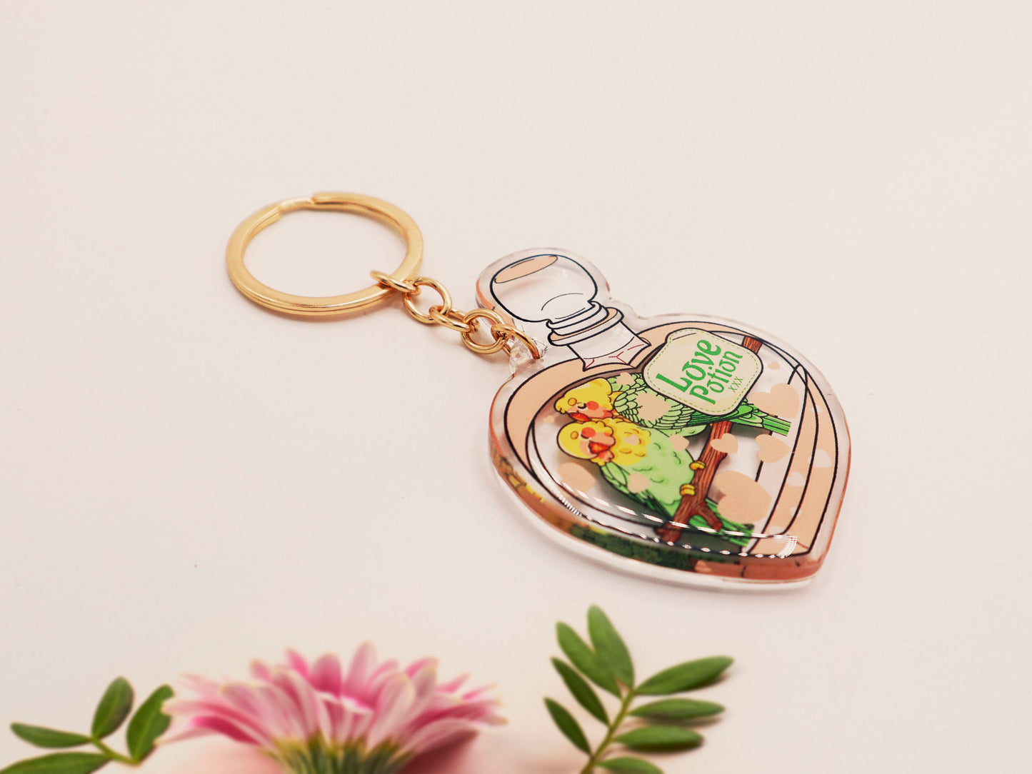 Clear acrylic double sided keyring with gold clasp, with a cute cartoon illustrated design of a heart shaped potion bottle containing two green and yellow love birds kissing each other labelled love potion xxx