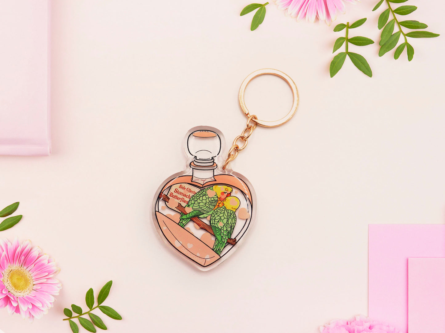 Clear acrylic double sided keyring with gold clasp, with a cute cartoon illustrated design of a heart shaped potion bottle containing two green and yellow love birds kissing each other labelled love potion xxx