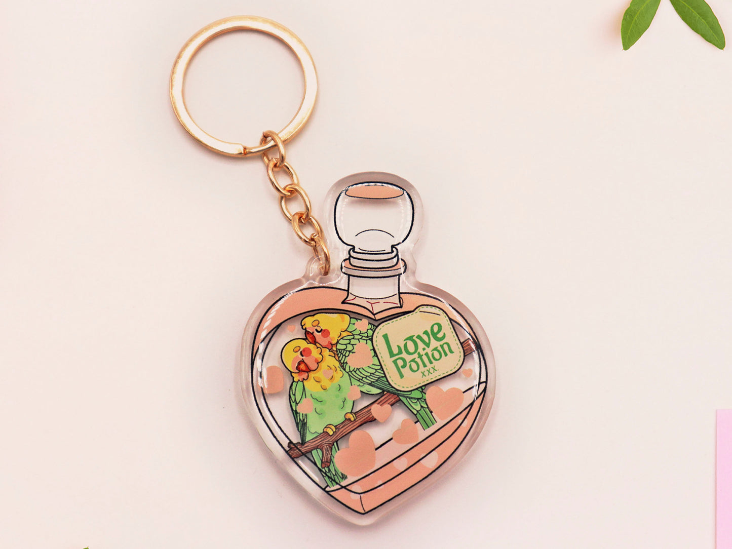 Clear acrylic double sided keyring with gold clasp, with a cute cartoon illustrated design of a heart shaped potion bottle containing two green and yellow love birds kissing each other labelled love potion xxx