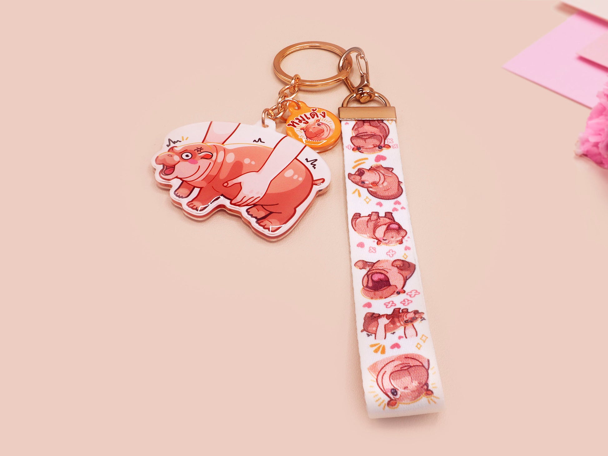 Acrylic keychain with a large charm of the fat angry pygmy hippo being picked up, a smaller charm of a cute baby hippo with the name Moo Deng in Thai and a matching mini lanyard of the pygmy hippo in multiple cute poses and expressions. 