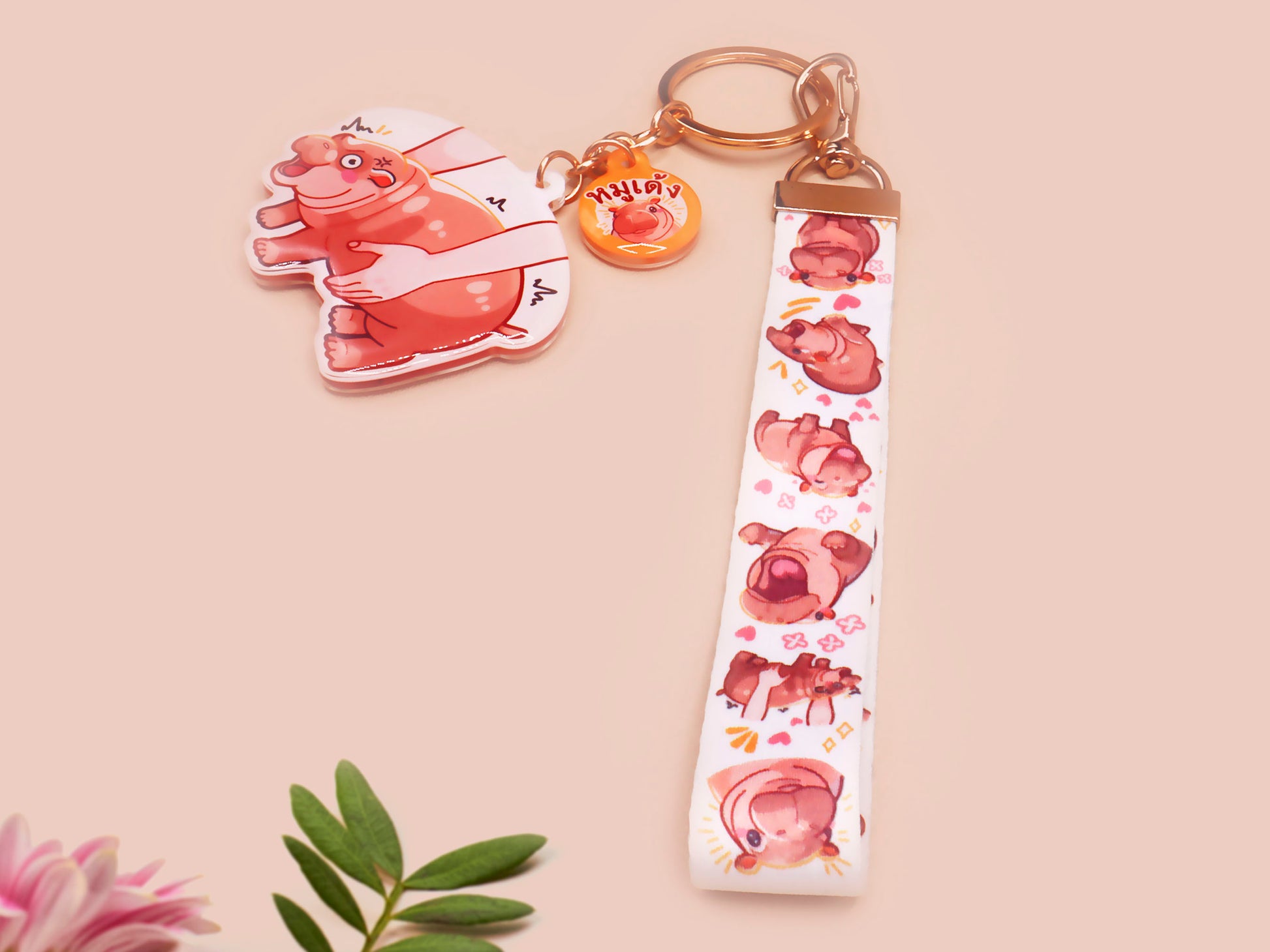 Acrylic keychain with a large charm of the fat angry pygmy hippo being picked up, a smaller charm of a cute baby hippo with the name Moo Deng in Thai and a matching mini lanyard of the pygmy hippo in multiple cute poses and expressions. 