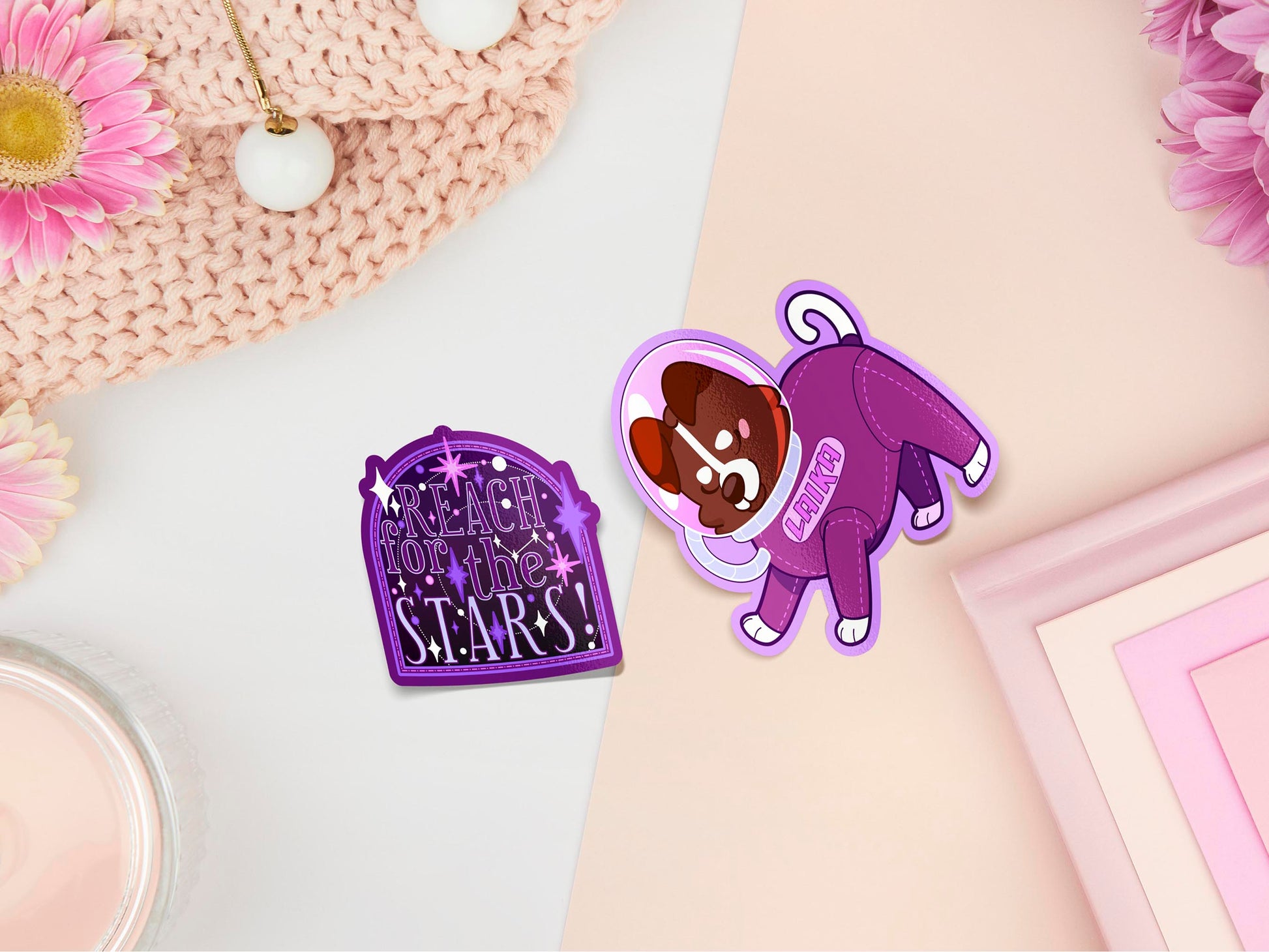 Two stickers of digitally illustrated cartoons, is the quote reach for the stars, surrounded by stars. The other is the Russian jack Russel space dog Laika in a purple space suit 