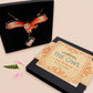 Mixed material handmade necklace of chibi cartoon flying barn owl with pearlescent wings carrying a removeable spell book charm, with a gold chain and black gift box with a yellow familiars collection gift sleeve.