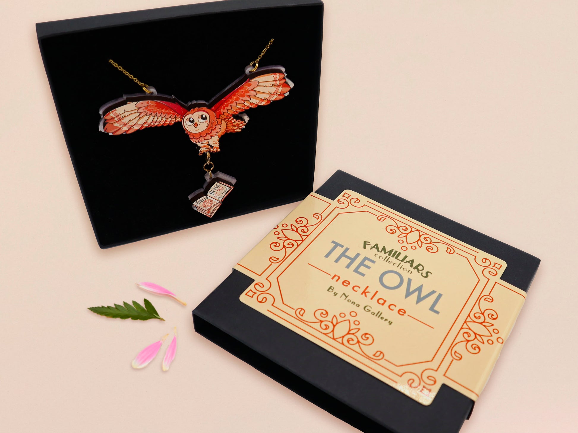 Mixed material handmade necklace of chibi cartoon flying barn owl with pearlescent wings carrying a removeable spell book charm, with a gold chain and black gift box with a yellow familiars collection gift sleeve.