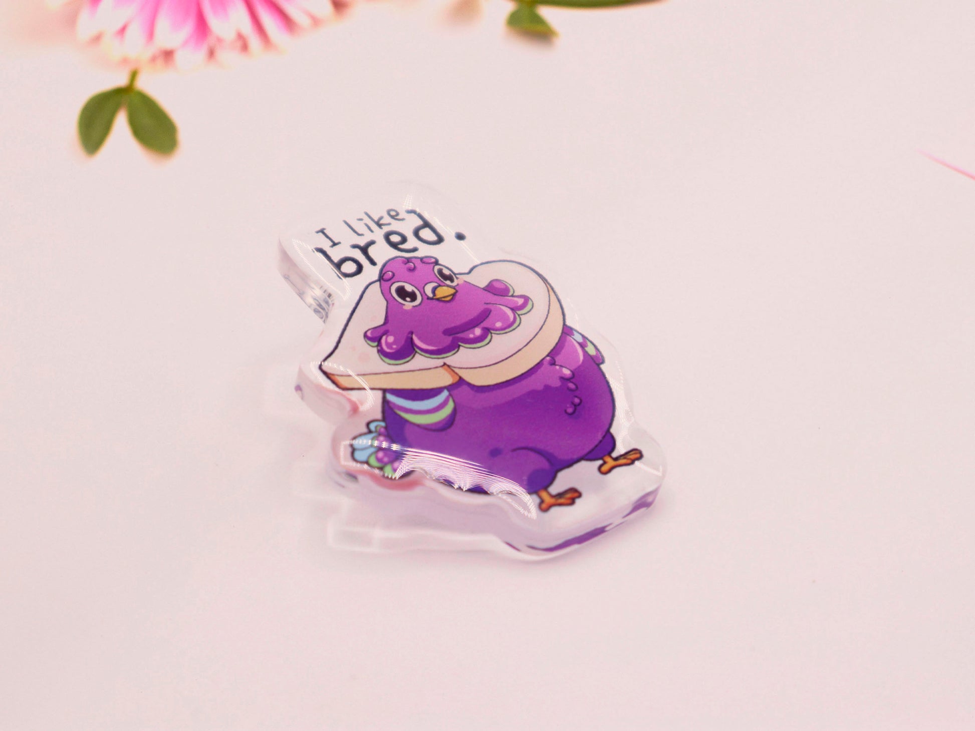An acrylic pin badge of a fat cartoon pigeon with a slice of bread stuck around their neck. Above is written the quote I like bred.