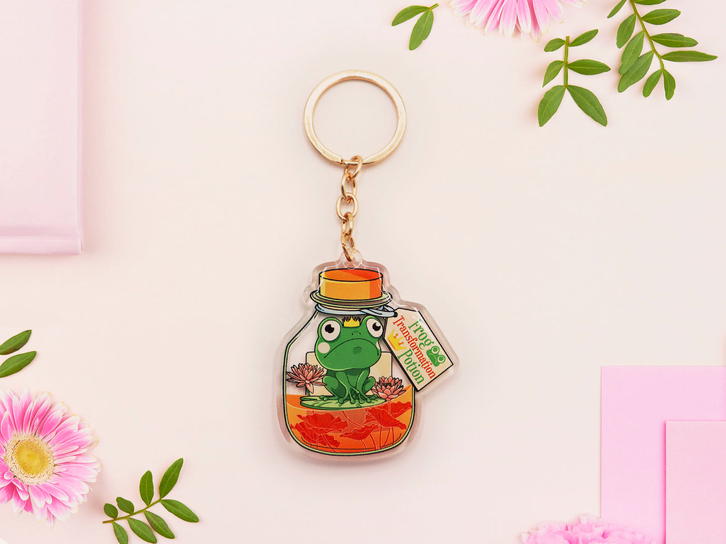 Double sided epoxy keychain with golden clasp of a frog inside a potion bottle looking glum wearing a small crown and sat on a lily pad., with the potion bottle labelled Frog Transformation Potion