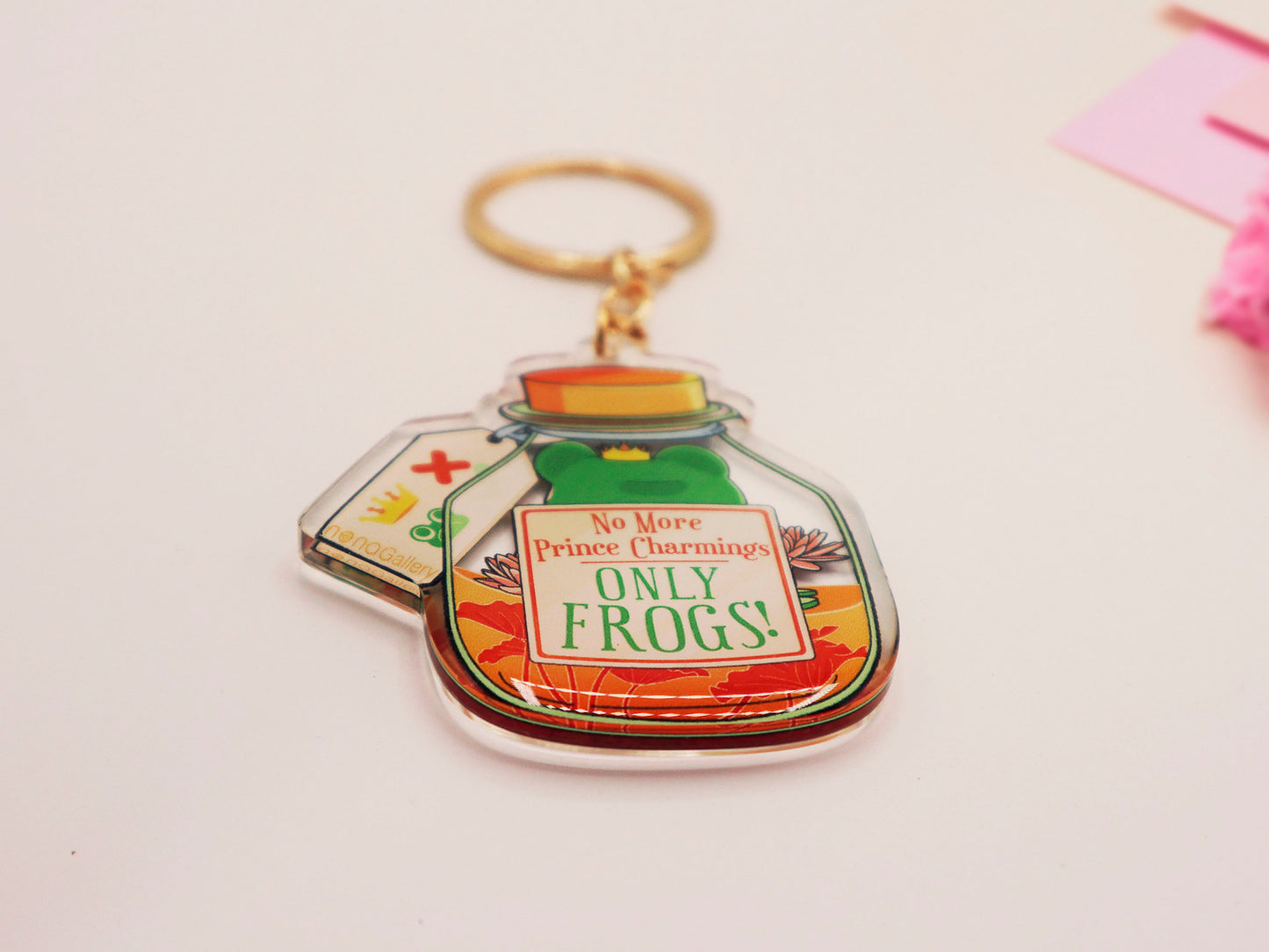 Double sided epoxy keychain with golden clasp of a frog inside a potion bottle looking glum wearing a small crown and sat on a lily pad., with the potion bottle labelled Frog Transformation Potion