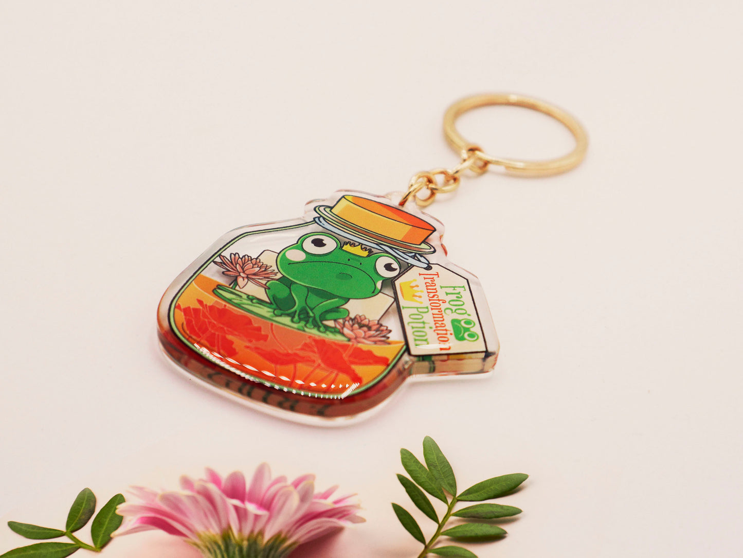 Double sided epoxy keychain with golden clasp of a frog inside a potion bottle looking glum wearing a small crown and sat on a lily pad., with the potion bottle labelled Frog Transformation Potion