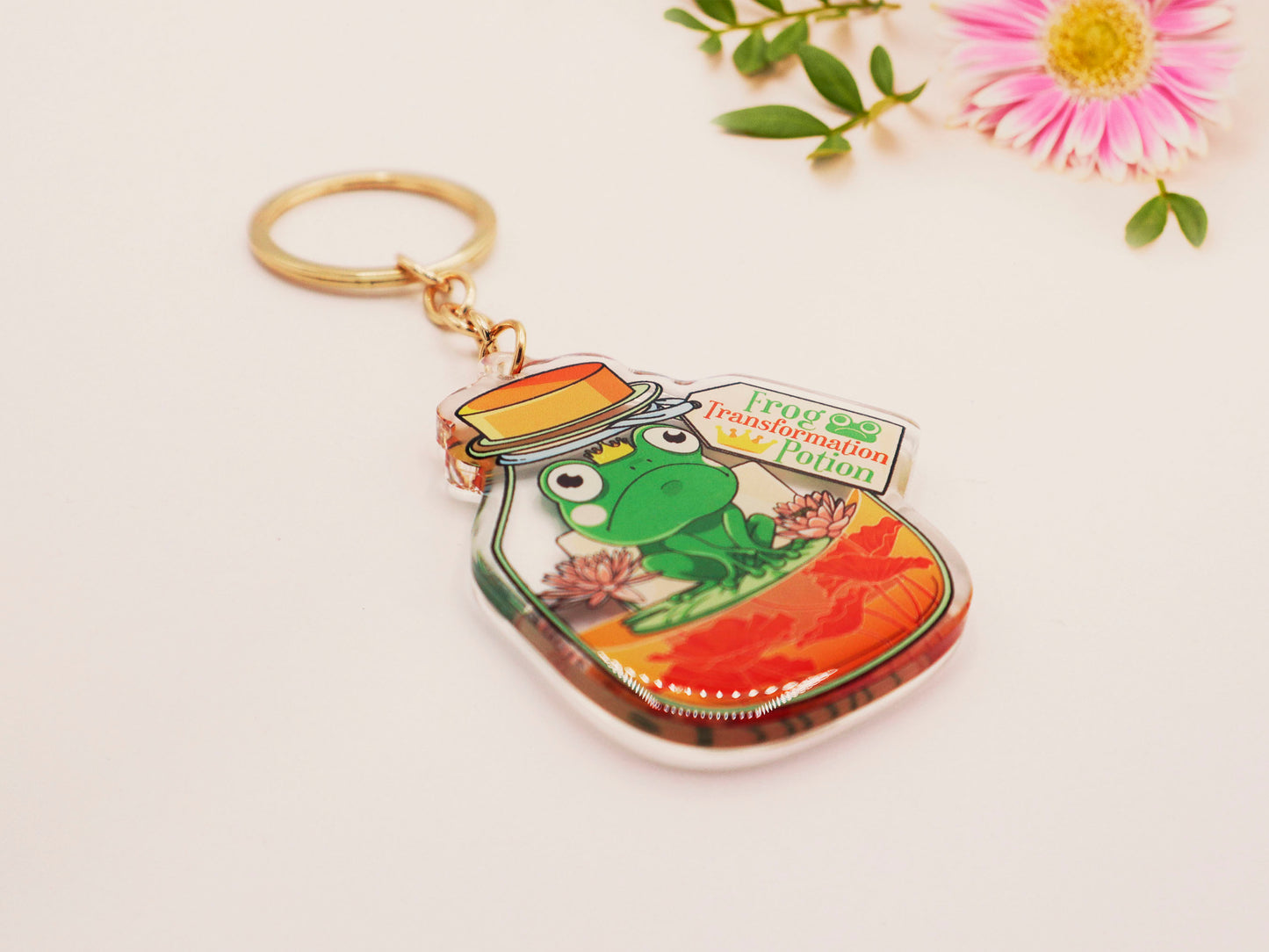 Double sided epoxy keychain with golden clasp of a frog inside a potion bottle looking glum wearing a small crown and sat on a lily pad., with the potion bottle labelled Frog Transformation Potion