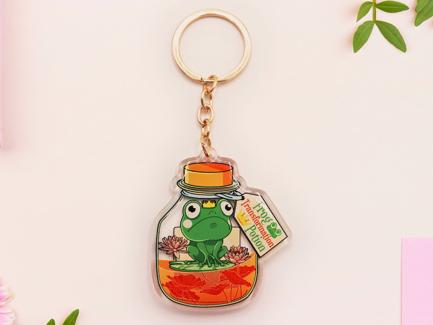 Double sided epoxy keychain with golden clasp of a frog inside a potion bottle looking glum wearing a small crown and sat on a lily pad., with the potion bottle labelled Frog Transformation Potion