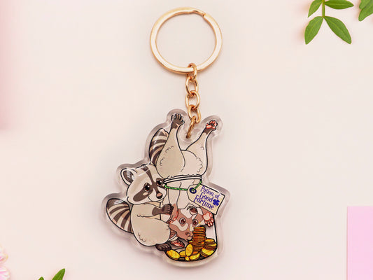 Clear acrylic keychain with gold chain clasp of two cheeky raccoons stealing gold coins from inside a potion bottle labelled potion of good fortune