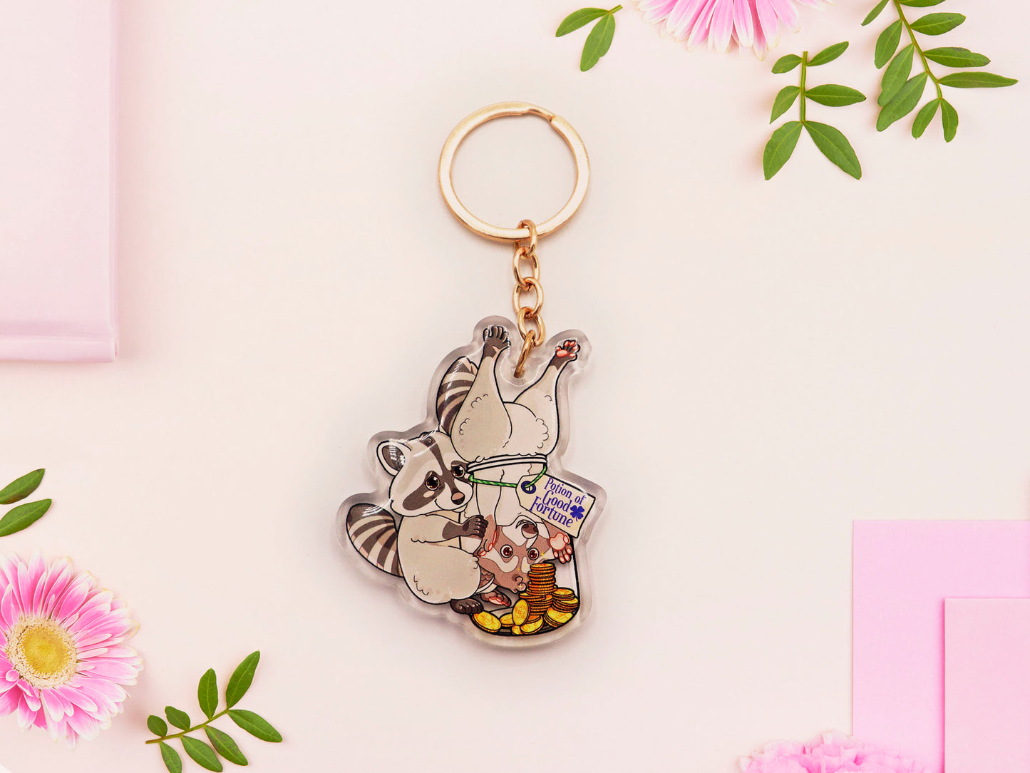 Clear acrylic keychain with gold chain clasp of two cheeky raccoons stealing gold coins from inside a potion bottle labelled potion of good fortune
