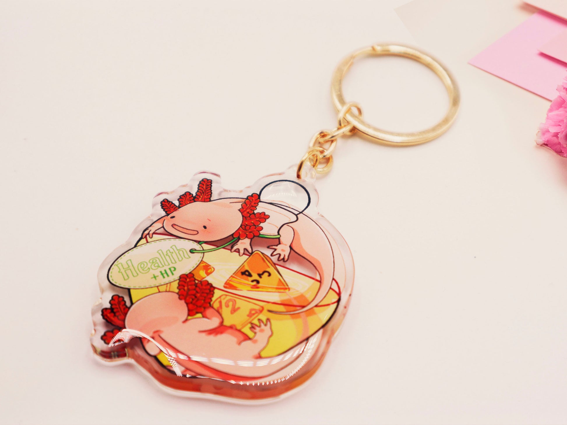 Clear acrylic double sided gold clasp keychain with a cartoon illustrated design of two pink axolotls on a potion bottle labelled health +hp and with a yellow liquid and four orange d4 dice inside.