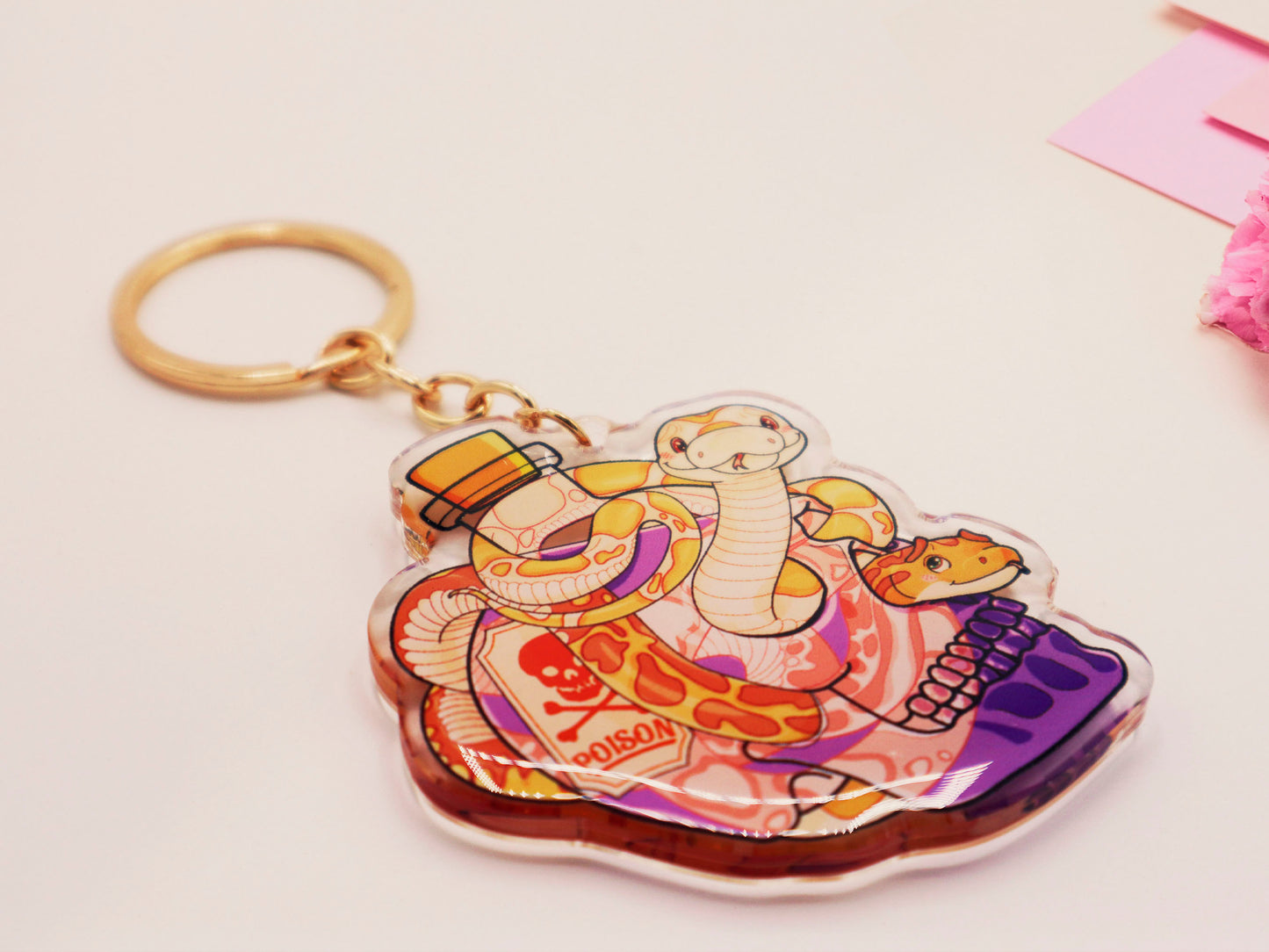 Clear acrylic double sided keyring with a cute cartoon illustrated skull shaped potion bottle with two yellow and brown corn snakes, filled with a purple liquid labelled with a skull and bones and the word poison.