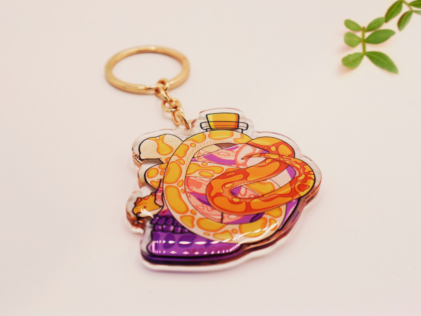 Clear acrylic double sided keyring with a cute cartoon illustrated skull shaped potion bottle with two yellow and brown corn snakes, filled with a purple liquid labelled with a skull and bones and the word poison.