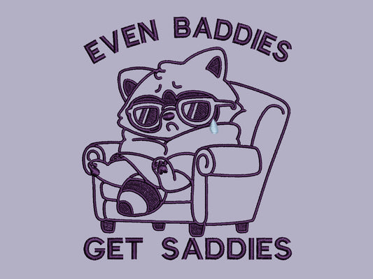 Purple embroidered cute design of a chibi raccoon crying wrapped in a blanket sat on a sofa with the quote Even Baddies Get Saddies