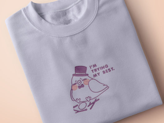 An embroidered purple t-shirt design of a cute pigeon wearing a top hat building a pathetic nest and the text I'm trying my best.