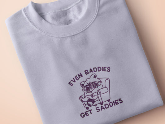 Purple embroidered cute t-shirt design of a chibi raccoon crying wrapped in a blanket sat on a sofa with the quote Even Baddies Get Saddies