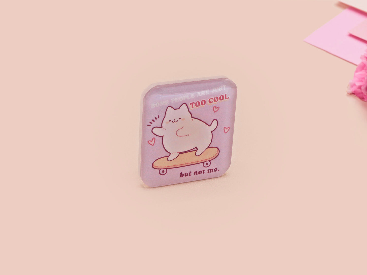 Clear acrylic pin badge with a pink heart-shaped rubber pin back clasp. The design of the pin badge is a cute chibi white cat skateboarding with the quote some people are just too cool but not me.