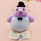 An adorable purple and blue pigeon bird plushie toy with cute embroidered eyes and facial expression, wearing a black top hat and bow tie, with a hang tag describing the pigeon as Susan.