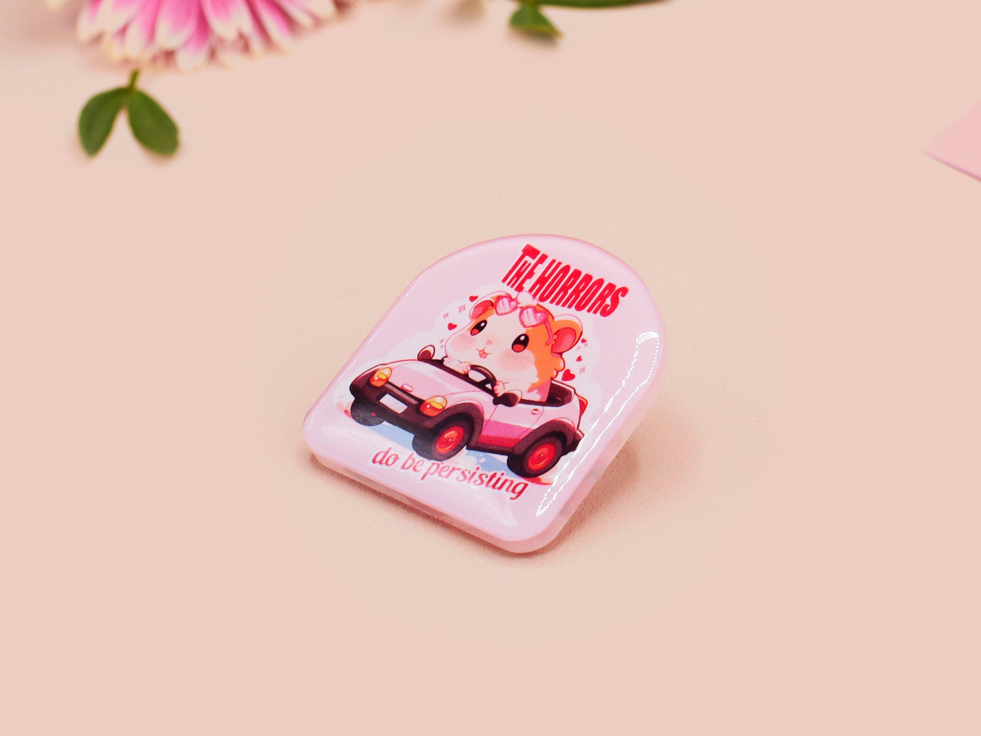Clear acrylic pin badge with a pink heart-shaped rubber pin back clasp. The design of the pin badge is a cute chibi guinea pig hamster driving a pink car and the quote the horrors do be persisting.