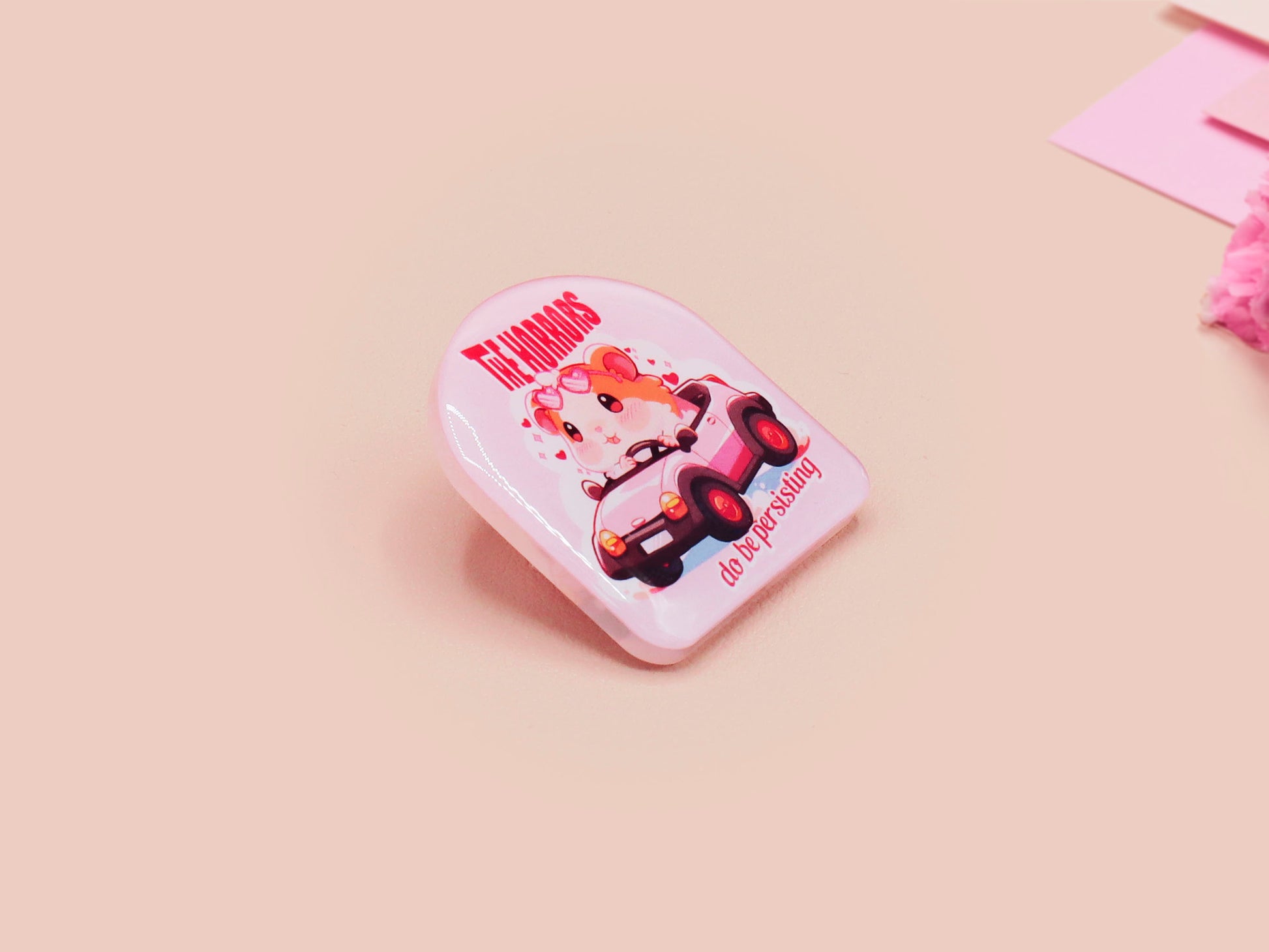 Clear acrylic pin badge with a pink heart-shaped rubber pin back clasp. The design of the pin badge is a cute chibi guinea pig hamster driving a pink car and the quote the horrors do be persisting.