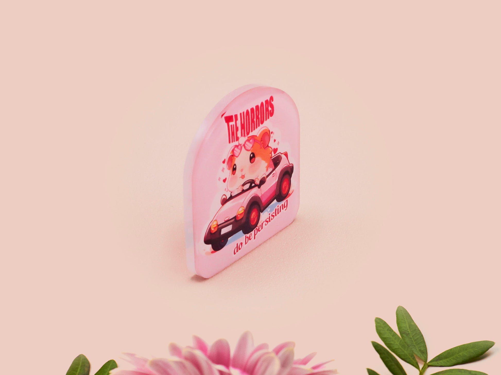 Clear acrylic pin badge with a pink heart-shaped rubber pin back clasp. The design of the pin badge is a cute chibi guinea pig hamster driving a pink car and the quote the horrors do be persisting.