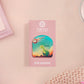 A clear acrylic pin badge with a pink heart-shaped pin back clasp. The design of the pin badge is of a cute long neck dinosur looking at an incoming asteroid with the quote uh oh.