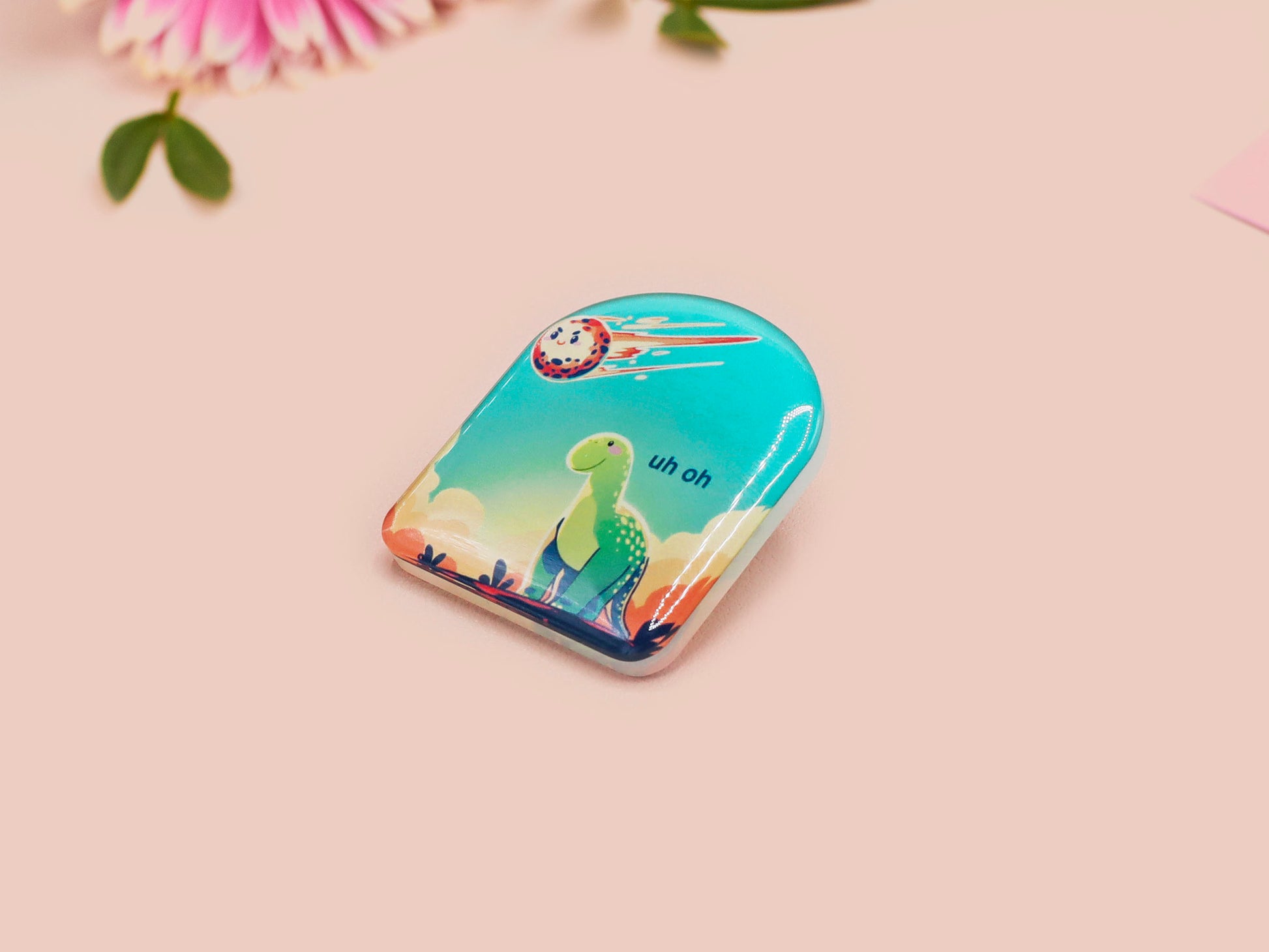 A clear acrylic pin badge with a pink heart-shaped pin back clasp. The design of the pin badge is of a cute long neck dinosur looking at an incoming asteroid with the quote uh oh.