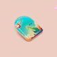A clear acrylic pin badge with a pink heart-shaped pin back clasp. The design of the pin badge is of a cute long neck dinosur looking at an incoming asteroid with the quote uh oh.