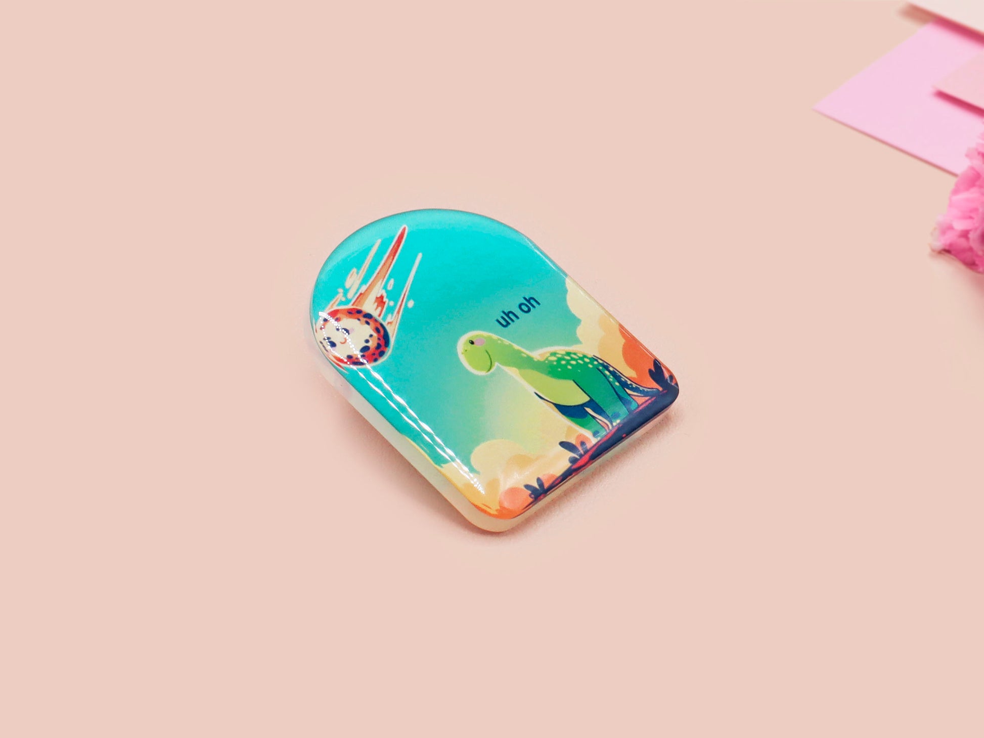 A clear acrylic pin badge with a pink heart-shaped pin back clasp. The design of the pin badge is of a cute long neck dinosur looking at an incoming asteroid with the quote uh oh.