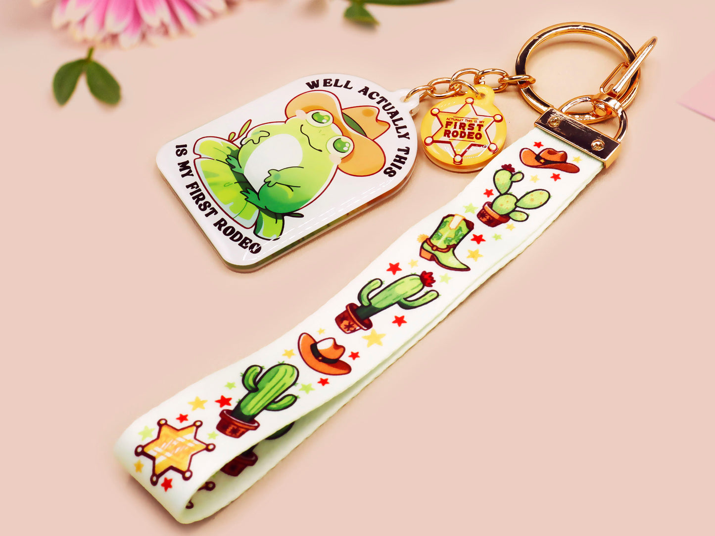 Acrylic epoxy cowboy frog keychain with a large acrylic charm of a cute cowboy frog wearing a stetson hat with the quote well actually this is my first rodeo, a smaller charm of a cute sherrifs badge and a matching cactus and cowboy boots and hats lanyard.