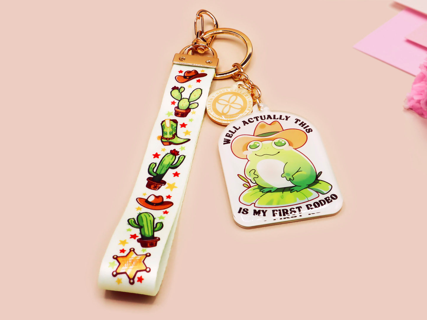 Acrylic epoxy cowboy frog keychain with a large acrylic charm of a cute cowboy frog wearing a stetson hat with the quote well actually this is my first rodeo, a smaller charm of a cute sherrifs badge and a matching cactus and cowboy boots and hats lanyard.