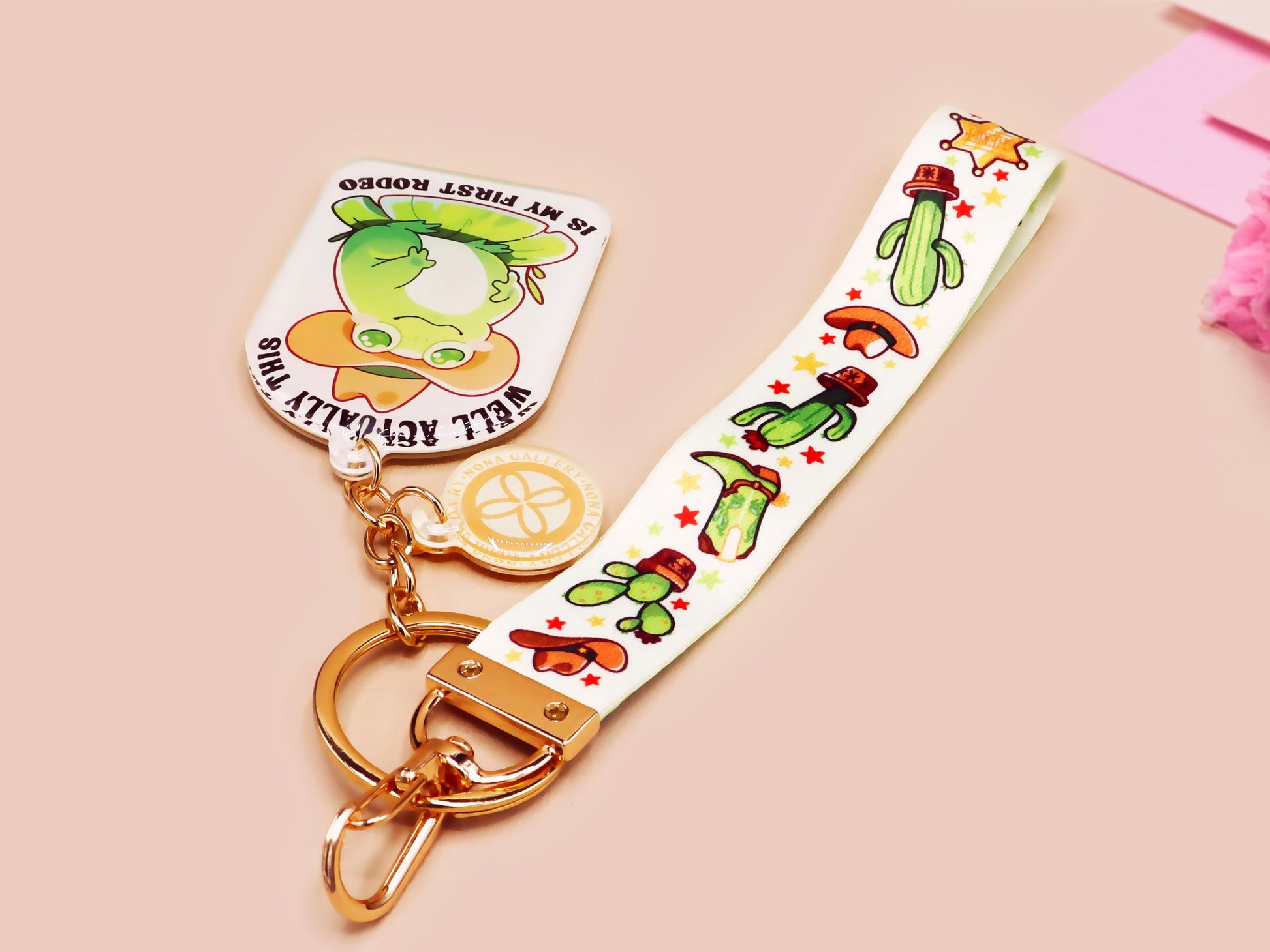 Acrylic epoxy cowboy frog keychain with a large acrylic charm of a cute cowboy frog wearing a stetson hat with the quote well actually this is my first rodeo, a smaller charm of a cute sherrifs badge and a matching cactus and cowboy boots and hats lanyard.
