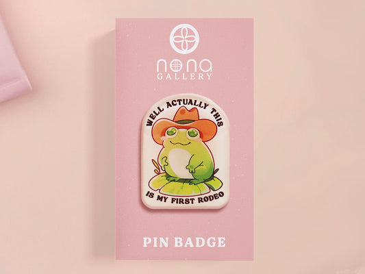 Clear acrylic pin badge with a pink heart-shaped rubber pin back clasp. The design of the pin badge is a cute chibi frog wearing a cowboy hat stetson and the quote well actually this is my first rodeo.