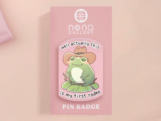 Clear acrylic pin badge with a pink heart-shaped rubber pin back clasp. The design of the pin badge is a cute chibi frog wearing a cowboy hat stetson and the quote well actually this is my first rodeo.