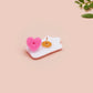 A clear acrylic pin badge with a pink heart-shaped pin back clasp. The design of the pin badge is of a cute chibi goose wearing a paper hat with the quote you silly goose.