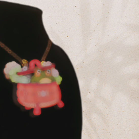 Video of front and back of mixed material handmade necklace of chibi cartoon frog sat inside a bubbling pearlescent witch's cauldron, with a gold chain and black gift box with a green familiars collection gift sleeve.